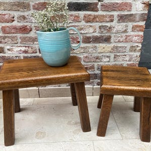 Puede incluir: Dos pequeños taburetes de madera hechos a mano con un acabado natural. Un taburete sostiene una jarra de cerámica azul claro llena de flores blancas. Los taburetes están colocados contra una pared de ladrillo.