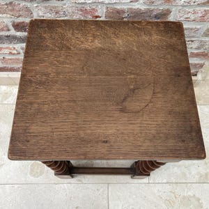 May include: A small, square wooden table with a dark brown finish. The tabletop has visible wood grain and a slightly worn appearance. The legs feature a decorative, turned design. The table is set against a brick wall and tiled floor.