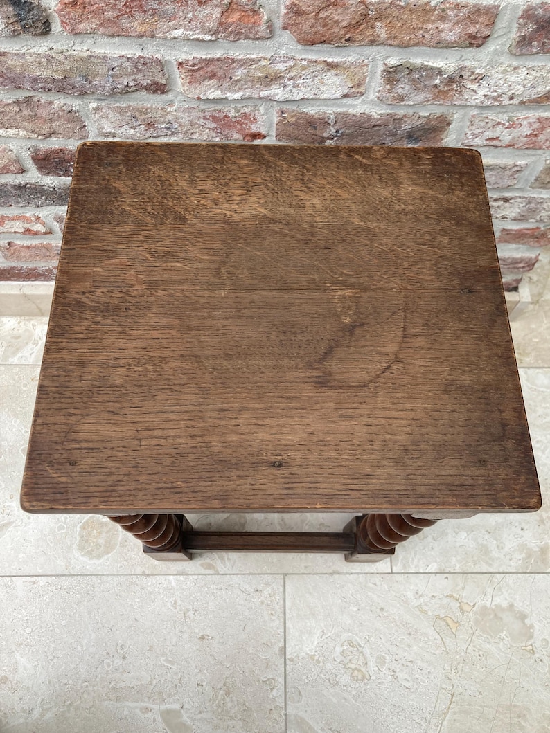 May include: A small, square wooden table with a dark brown finish. The tabletop has visible wood grain and a slightly worn appearance. The legs feature a decorative, turned design. The table is set against a brick wall and tiled floor.