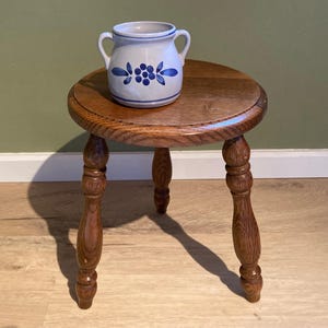 May include: A small, round wooden stool with three turned legs. A ceramic jug with two handles and blue floral designs sits on top. The stool and jug are set against a green wall and wooden floor.