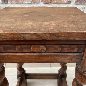 May include: A small, square, antique wooden table with a dark brown finish. The table features a carved apron and intricately turned legs. The tabletop is made of solid wood, showing the natural grain. The table is approximately 20 inches tall.