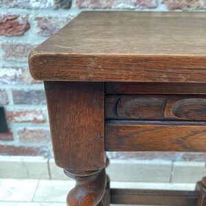 May include: A close-up of a small, antique wooden table. The table has a rectangular top and ornate, turned legs. The wood has a warm, brown tone with visible grain and some wear, suggesting age and use. The table is likely a side table or small accent piece.