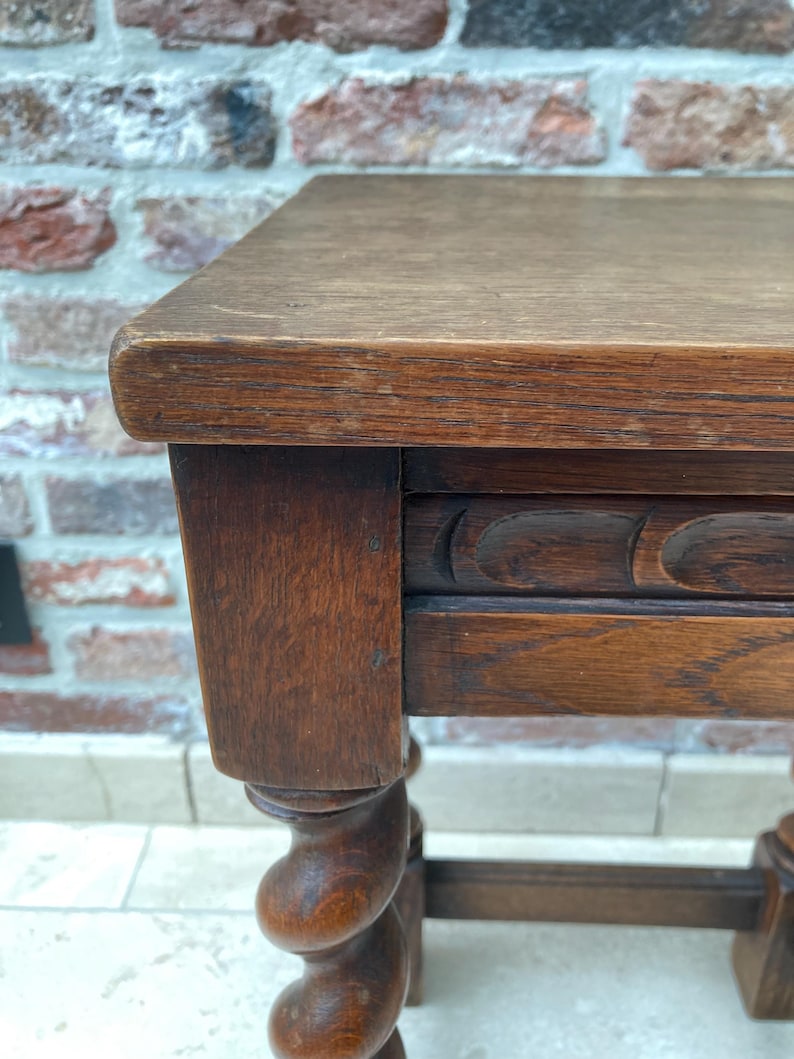 May include: A close-up of a small, antique wooden table. The table has a rectangular top and ornate, turned legs. The wood has a warm, brown tone with visible grain and some wear, suggesting age and use. The table is likely a side table or small accent piece.