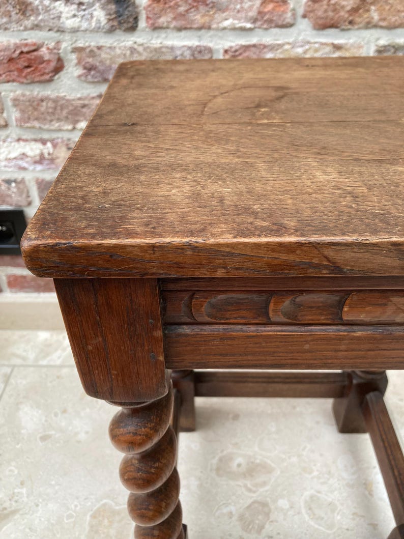May include: A close-up of a small, antique wooden table. The table has a rectangular top and a carved apron. One leg is a turned spiral design. The wood has a warm, brown tone and visible grain.