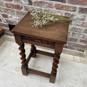 May include: A small, antique wooden stool with a square top and intricately carved details. The legs are turned in a spiral design. A sprig of white flowers rests on the top. The stool is made of dark brown wood.