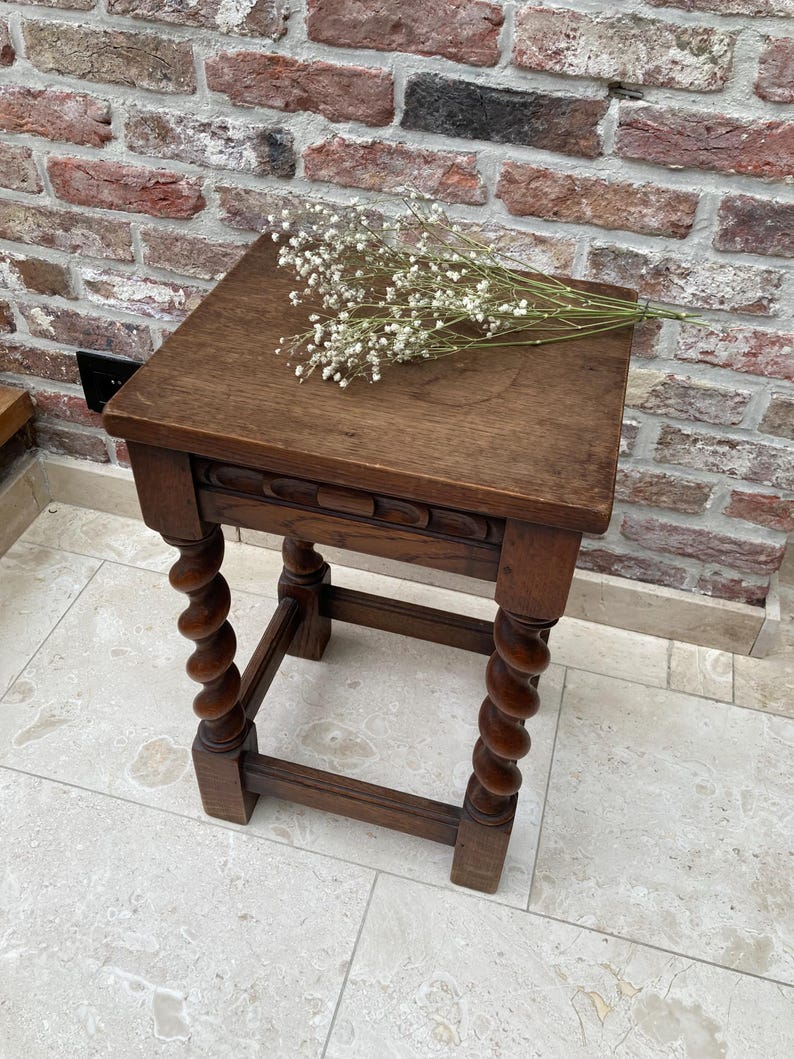 May include: A small, antique wooden stool with a square top and intricately carved details. The legs are turned in a spiral design. A sprig of white flowers rests on the top. The stool is made of dark brown wood.