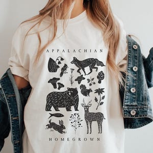 May include: Cream-colored t-shirt with a black graphic featuring Appalachian wildlife and flora. The design includes a bear, fox, deer, rabbit, and other elements, with the words "APPALACHIAN HOMEGROWN" printed above and below the illustrations.