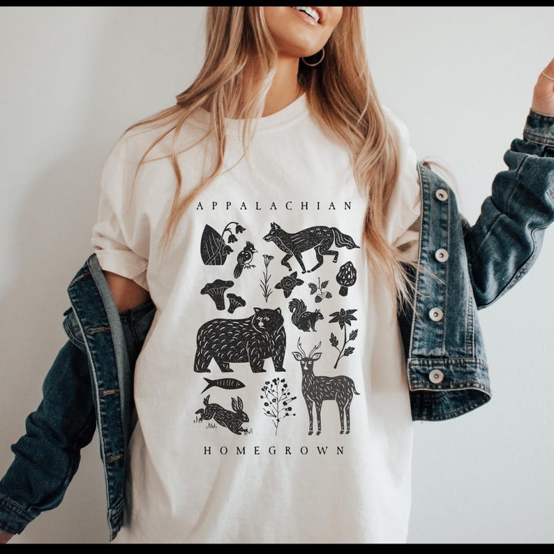 May include: Cream-colored t-shirt with a black graphic featuring Appalachian wildlife and flora. The design includes a bear, fox, deer, rabbit, and other elements, with the words "APPALACHIAN HOMEGROWN" printed above and below the illustrations.