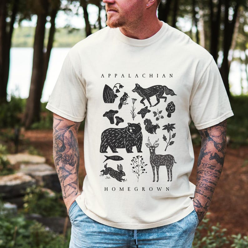May include: Cream-colored t-shirt featuring a black graphic design with the words "APPALACHIAN HOMEGROWN." The design includes illustrations of a bear, fox, deer, rabbit, mushrooms, and other natural elements.