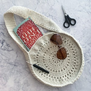 May include: Cream-colored, crocheted tote bag with a circular design. The bag contains a copy of "The Great Gatsby", sunglasses, and a crochet hook. A pair of black scissors rests nearby. The bag has a long strap for over-the-shoulder wear.
