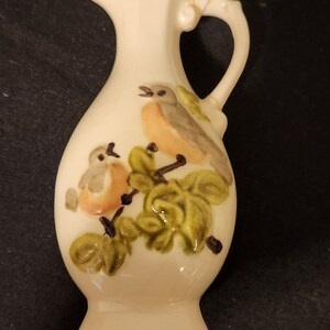 Könnte beinhalten: Ein kleiner weißer Keramik Krug mit Henkel. Der Krug hat ein Blumenmuster mit zwei Vögeln und grünen Blättern.