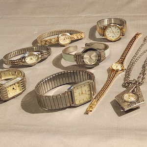 May include: A collection of various wristwatches and a pendant watch. The watches feature gold and silver tones, with different band styles including stretch, cuff, and link bands. The watch faces are primarily round or rectangular.