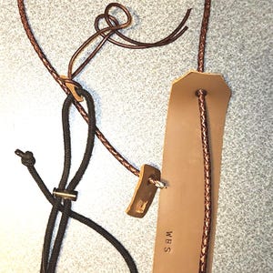 May include: A brown leather and braided cord lanyard with a rectangular leather tag. The tag has the initials "WBS" printed on it. The lanyard has a black cord with a metal clasp.