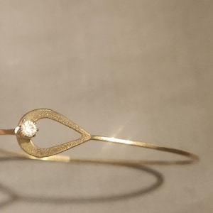 May include: A delicate gold-toned bracelet featuring a teardrop-shaped design with a sparkling, round, clear gemstone at the top. The bracelet has a thin, minimalist band, and the teardrop shape is open in the center.