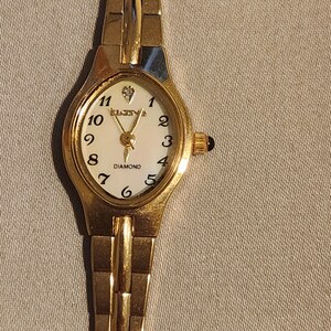 May include: A gold-toned Elgin watch with a white face and black numerals. The watch features a diamond accent at the 12 o'clock position and a gold-toned band. The watch is a classic design, suitable for everyday wear.