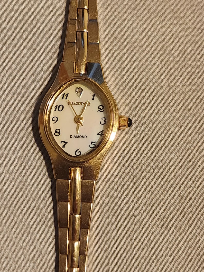 May include: A gold-toned Elgin watch with a white face and black numerals. The watch features a diamond accent at the 12 o'clock position and a gold-toned band. The watch is a classic design, suitable for everyday wear.