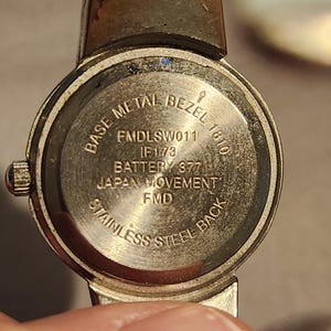 May include: Close-up of a vintage wristwatch back. The watch features a stainless steel back and a base metal bezel. Text on the watch includes "Base Metal Bezel", "Japan Movement", and "Stainless Steel Back". The watch has a silver and gold tone.