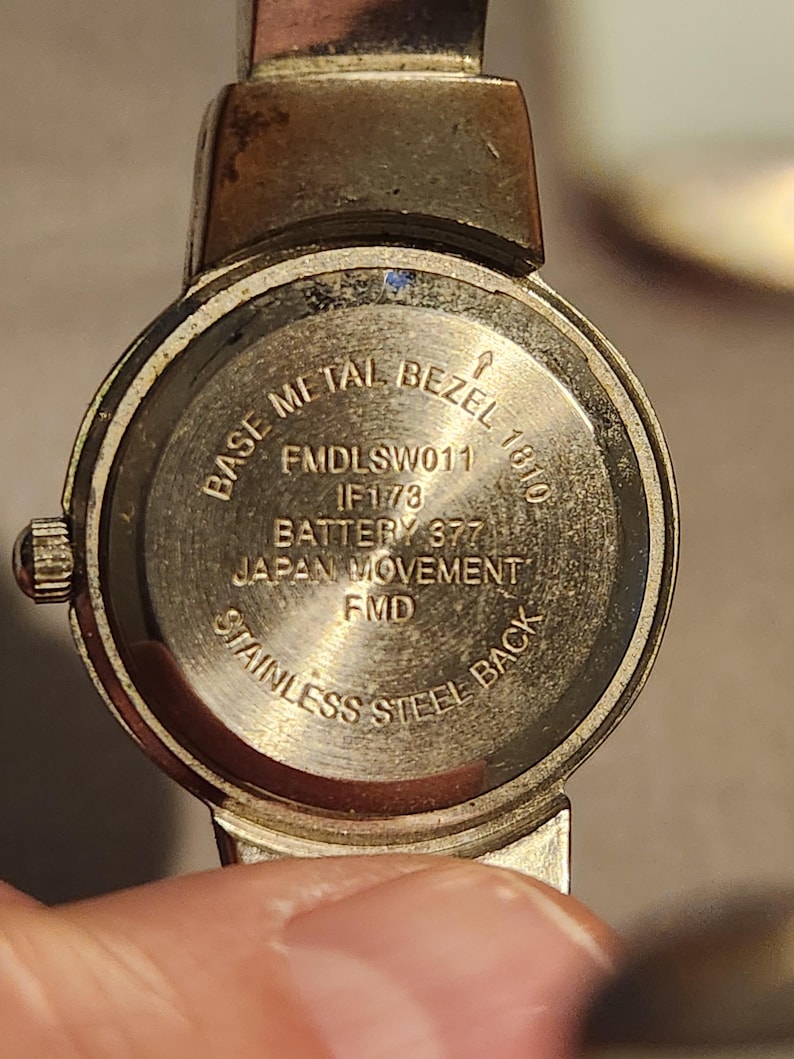 May include: Close-up of a vintage wristwatch back. The watch features a stainless steel back and a base metal bezel. Text on the watch includes "Base Metal Bezel", "Japan Movement", and "Stainless Steel Back". The watch has a silver and gold tone.