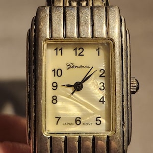 May include: Silver-toned Geneva watch with a rectangular face. The watch has a cream-colored dial with black numerals and hands. The watch band is also silver-toned. The words "Geneva" and "Japan Movt" are printed on the watch face.