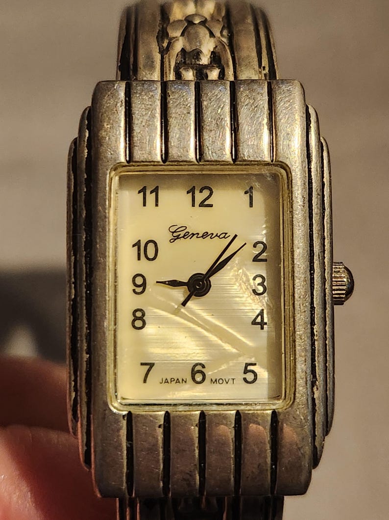 May include: Silver-toned Geneva watch with a rectangular face. The watch has a cream-colored dial with black numerals and hands. The watch band is also silver-toned. The words "Geneva" and "Japan Movt" are printed on the watch face.