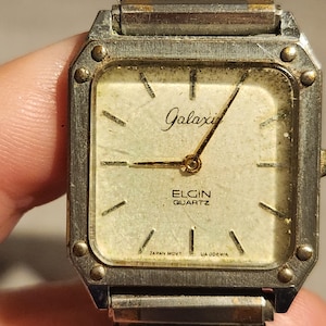 May include: Vintage Elgin Quartz watch with a square face. The watch has a silver-tone metal case and a cream-colored dial with gold-tone hands and markers. The watch face reads "Galaxi" and "Elgin Quartz". The watch band is silver-tone metal.