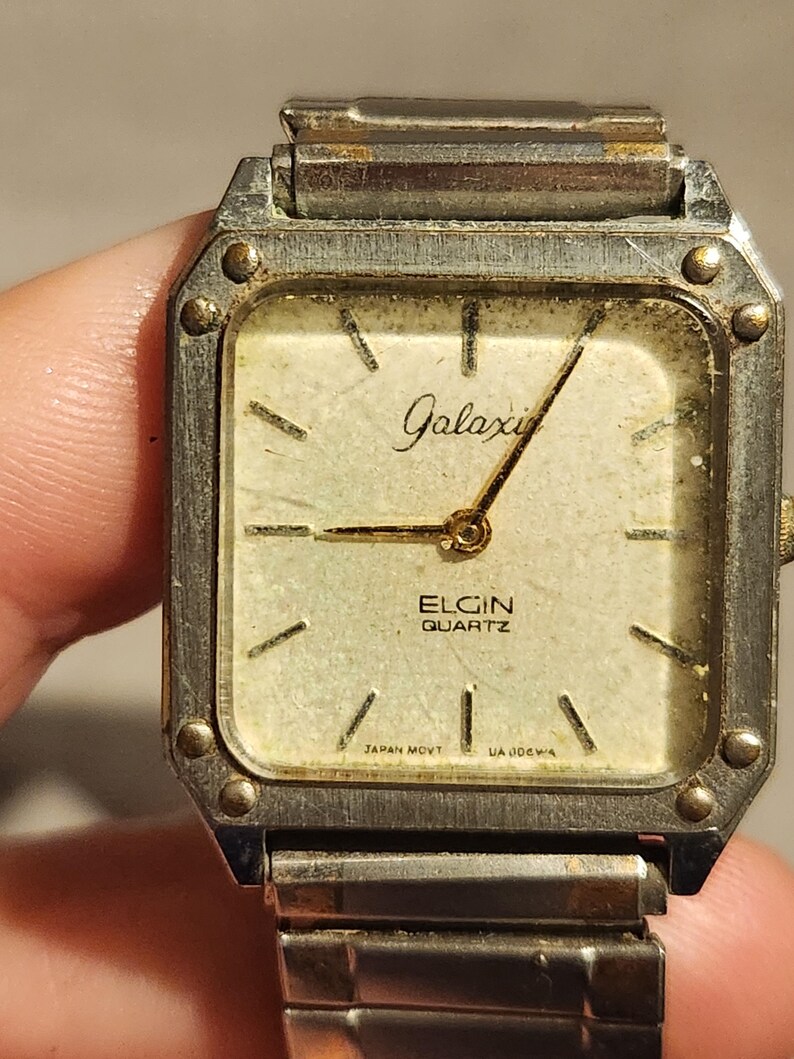 May include: Vintage Elgin Quartz watch with a square face. The watch has a silver-tone metal case and a cream-colored dial with gold-tone hands and markers. The watch face reads "Galaxi" and "Elgin Quartz". The watch band is silver-tone metal.