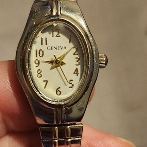 May include: Silver-toned Geneva watch with an oval face and a white dial. The watch features gold-colored hands and numerals, with the brand name "GENEVA" displayed. The watch band is a silver-toned, flexible, and expandable design.