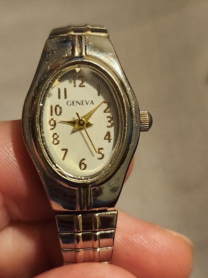 May include: Silver-toned Geneva watch with an oval face and a white dial. The watch features gold-colored hands and numerals, with the brand name "GENEVA" displayed. The watch band is a silver-toned, flexible, and expandable design.