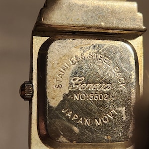 May include: Close-up of a vintage Geneva watch. The watch back is stainless steel and features the brand name, model number 5502, and "Japan Movt." The watch has a silver-toned metal band and case.