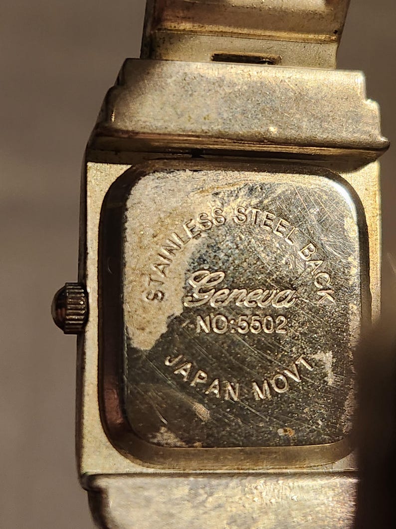 May include: Close-up of a vintage Geneva watch. The watch back is stainless steel and features the brand name, model number 5502, and "Japan Movt." The watch has a silver-toned metal band and case.