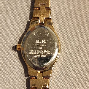May include: Gold-toned wristwatch with a rectangular face and a matching link band. The watch back is visible, displaying the text "ELL 10/ VC11-379 098 BASE METAL BEZEL STAINLESS STEEL BACK JAPAN MOVT."