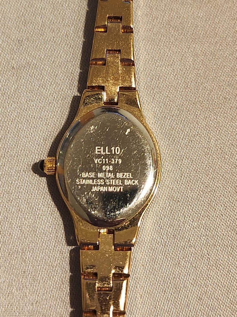 May include: Gold-toned wristwatch with a rectangular face and a matching link band. The watch back is visible, displaying the text "ELL 10/ VC11-379 098 BASE METAL BEZEL STAINLESS STEEL BACK JAPAN MOVT."