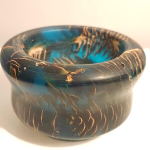 May include: A small, round bowl crafted from translucent blue resin with embedded dark brown, wood-like grain patterns. The vessel features a smooth, polished finish with a wide, rounded rim and a deep, concave interior that reveals the swirling colors.