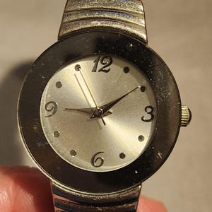May include: A silver-toned wristwatch with a round face featuring a silver dial, black bezel, and silver hands. The watch has a white band and is accented with silver metal. The numbers 3, 6, 9, and 12 are visible.