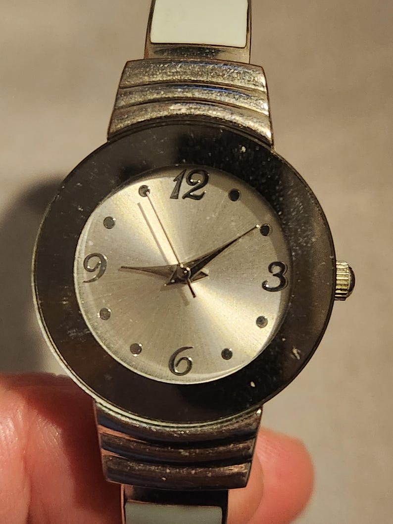 May include: A silver-toned wristwatch with a round face featuring a silver dial, black bezel, and silver hands. The watch has a white band and is accented with silver metal. The numbers 3, 6, 9, and 12 are visible.