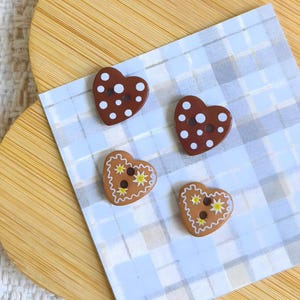 Peut inclure: Quatre boutons en forme de cœur. Deux sont marron avec des pois blancs, et deux sont marron clair avec des motifs d'étoiles jaunes. Ils reposent sur un papier à carreaux bleu et blanc, sur une surface en forme de cœur en bois.