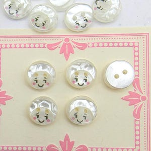 Può includere: Una collezione di bottoni bianchi perlati con un design a faccina sorridente. I bottoni sono disposti su una scheda decorativa con un bordo rosa. I bottoni hanno due fori per cucire.