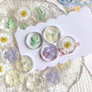 May include: A collection of circular clear resin buttons featuring embedded pressed flowers including white daisies, blue forget-me-nots, yellow blossoms, and green foliage. Some buttons are attached to a white scalloped display card with text.