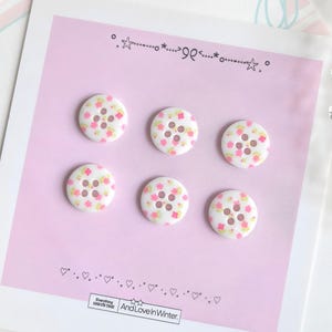 May include: Six round white buttons featuring a small pink and yellow floral pattern, each with four center holes. The buttons are displayed on a light pink card with decorative star and heart borders and the text And Love In Winter.
