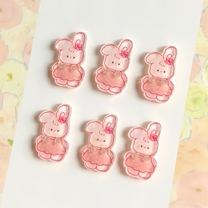 May include: Six small, translucent acrylic charms shaped like cartoon rabbits. Each rabbit is light pink with a darker pink dress, a small flower detail on the head, and a simple facial expression. The charms are arranged in two rows of three.