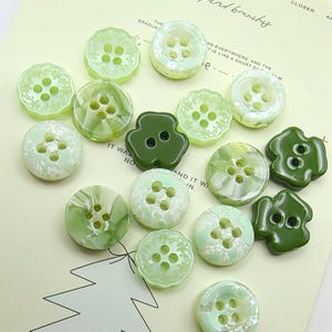 Può includere: Una collezione di bottoni verdi in varie forme e tonalità, inclusi design rotondi e a forma di trifoglio. Alcuni bottoni hanno una finitura perlata, mentre altri sono in tinta unita. I bottoni sono disposti su carta verde chiaro con testo scritto a mano.