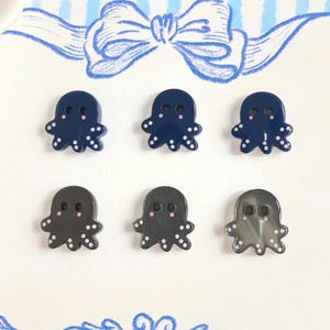 Puede incluir: Seis botones pequeños con forma de pulpo dispuestos en dos filas. La fila superior presenta tres botones azul oscuro con puntos blancos y detalles rosas. La fila inferior contiene tres botones gris oscuro, ligeramente translúcidos.