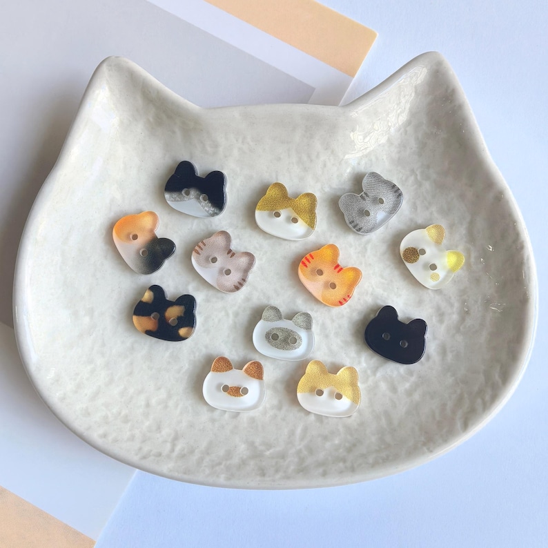 May include: A collection of small, two-hole buttons shaped like cat heads. The buttons feature various colors and patterns, including solid black, tortoiseshell, orange tabby, grey striped, and white with gold accents, all crafted from a smooth material.