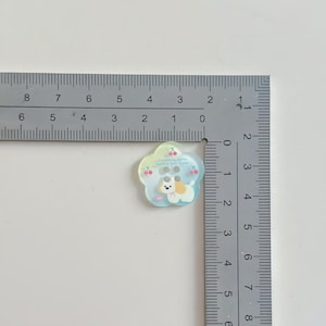 May include: A flower-shaped button with a cute dog illustration, measuring approximately 1 inch in diameter. The button has a light blue and yellow gradient, with cherry and text details. It features two buttonholes for sewing.