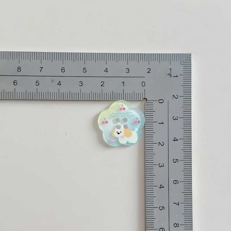 May include: A flower-shaped button with a cute dog illustration, measuring approximately 1 inch in diameter. The button has a light blue and yellow gradient, with cherry and text details. It features two buttonholes for sewing.