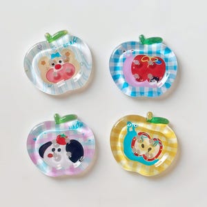 May include: Four decorative apple-shaped coasters. Each coaster features a different design: a bear, a red apple with a crescent moon, a dog with a strawberry, and a snail. The coasters have a clear, glossy finish with colorful illustrations.