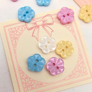 May include: A collection of flower-shaped buttons in various pastel colors, including blue, pink, yellow, and white. Each button has two holes for sewing. The buttons are arranged on a light-colored card with a bow illustration.
