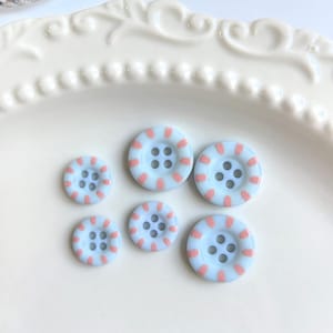 May include: Six light blue buttons with pink striped accents. The buttons have four holes each and are arranged on a white surface. These decorative buttons are ideal for sewing and craft projects.