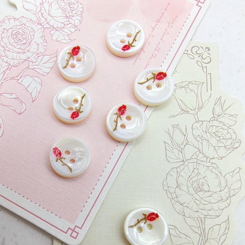 May include: A collection of white, round buttons with a pearlescent sheen. Each button features a hand-painted red rose with green stem and gold accents. The buttons have two or three holes for sewing and are arranged on a pink and cream background.