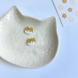 May include: Two small, cat-shaped buttons with a clear base and a gold-colored top section. Each button features two holes for attachment. They are resting inside a white, textured, cat-shaped ceramic dish next to a card with orange flower icons.